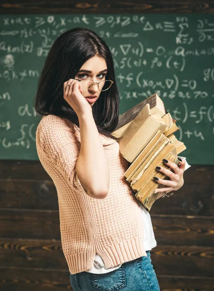 Chica morena en cárdigan rosa mirando por encima de sus gafas. Vista lateral hermosa chica universitaria llevando pila de libros antiguos — Foto de Stock