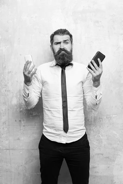 manager or bearded man with long beard and hair on misunderstanding face in tie and white shirt on beige background compare mobile phone and smartphone, conversation and information, businessman