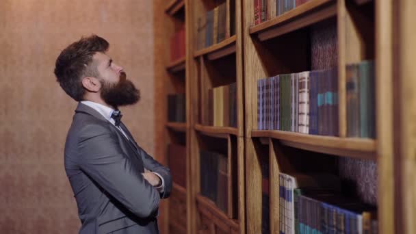 Человек в костюме стоит перед книжным шкафом в библиотеке и выбирает книгу. Концепция библиотеки . — стоковое видео