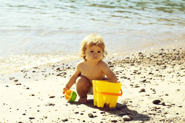 Piccolo curioso divertente bambino biondo seduto sulla spiaggia della costa del mare con acqua ondulata — Foto Stock