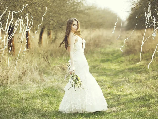 Hübsche Braut im Freien. schöne junge Braut in weißem Brautkleid mit Blumen im Feld mit Bändern an Bäumen im Freien — Stockfoto