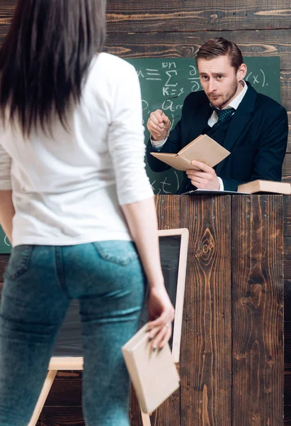 Γυρίστε πίσω μελαχρινή κοπέλα κρατώντας το βιβλίο στο χέρι της. Αυστηρός δάσκαλος βλέπει του φοιτήτριας ενώ στέκεται στο ρόστρου — Φωτογραφία Αρχείου