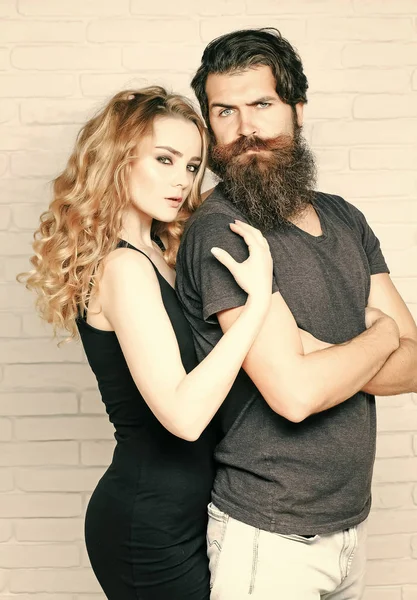 Hipsterism, subculture, trend. Man with beard and woman with long blond hair. Fashion, beauty, style concept. Girl and bearded hipster. Couple in love hug on white brick wall.