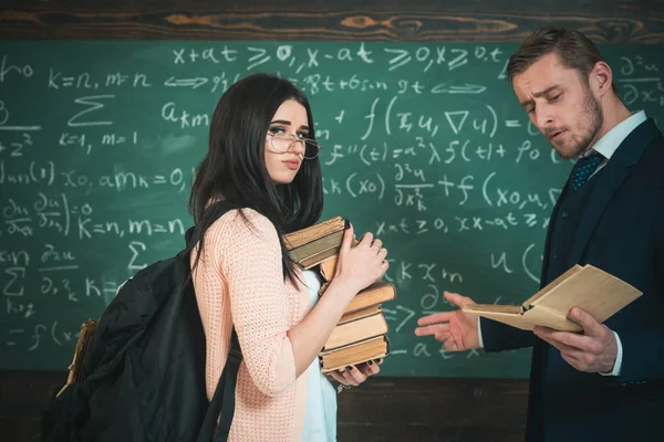 교수를 읽는 동안 책의 더미를 들고 안경을 쓰고 분홍색 카디 건 젊은 갈색 머리. 녹색 보드 앞에 서 있는 교사 및 대학 여자 — 스톡 사진