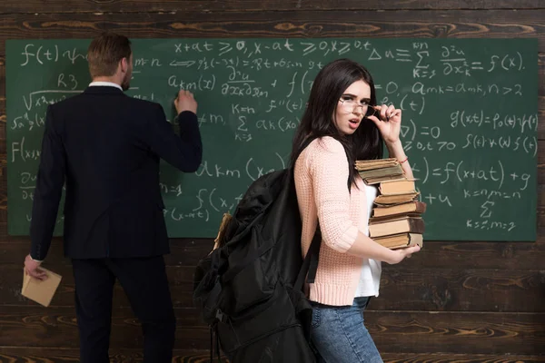 彼女は彼女が必要な本を持っています。女性学生は学校で黒板に書く先生男と本スタックでメガネとバックパックで見る。試験を準備します。知識に自信を持っています。勉強が大好き — ストック写真