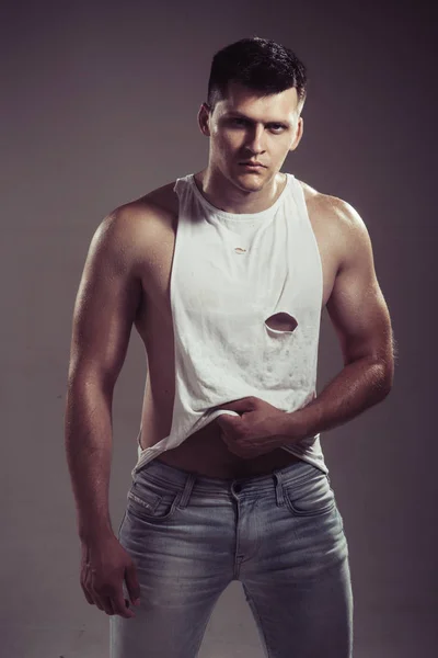 Hombre en camiseta sexy y jeans sobre fondo gris. Hombre con manos musculosas con bíceps y tríceps —  Fotos de Stock