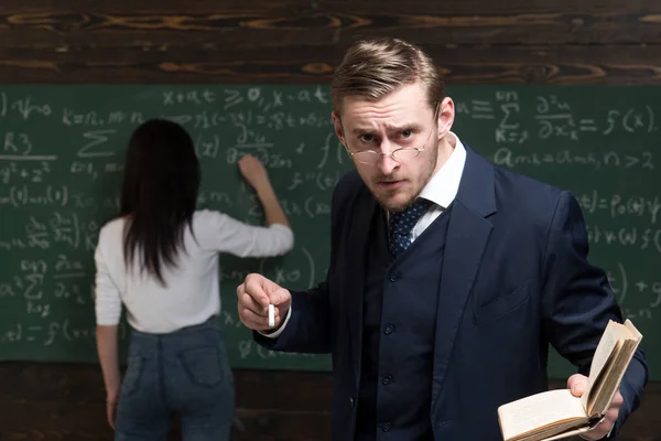 Der strenge Professor blickt Studenten über die Brille, während er Buch und Kreide in den Händen hält. Studentin schreibt auf grüne Tafel — Stockfoto