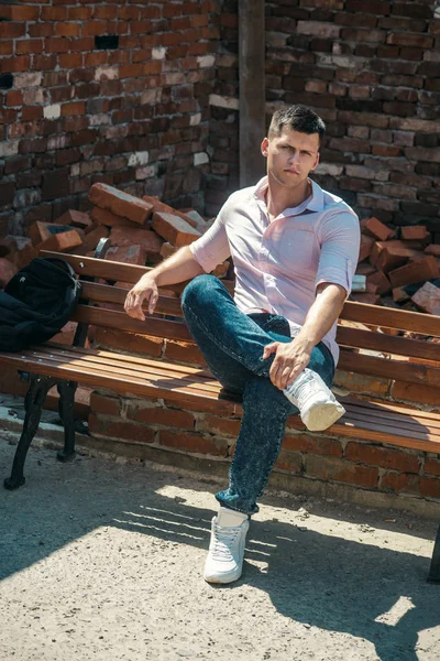 Homem relaxar no banco ao ar livre ensolarado. Macho na moda camisa e jeans moda. Modelo de moda em alvenaria de tijolo vermelho. Um tipo bonito com mochila de férias. Conceito de estilo jovem — Fotografia de Stock