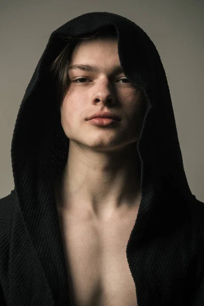 Tipo con cara confiada en capucha negra, fondo gris. Hombre con la piel lisa en la cara, cuello y pecho mirando a la cámara. Macho en albornoz negro con capucha, de cerca. Concepto de masculinidad . —  Fotos de Stock