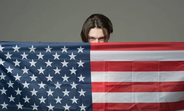 Un hombre con cara seria sostiene la bandera de EE.UU., escondiéndose detrás de la bandera. Un tipo escondido en EE. UU., América lo protege, fondo gris. Concepto de privacidad y conspiración . — Foto de Stock