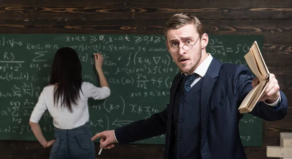 Agitated young professor teaching math at college. Brunette female student solving equation on green board — Stock Photo, Image