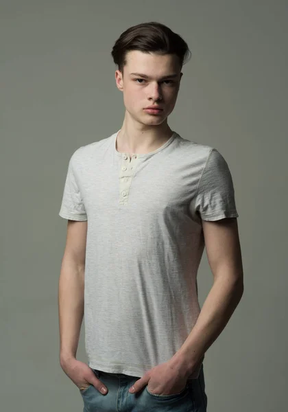 Portrait en studio d'un jeune homme brun sérieux aux cheveux de longueur moyenne isolé sur fond gris. Beau adolescent en T-shirt blanc et jean bleu rayonnant de confiance, concept de succès — Photo