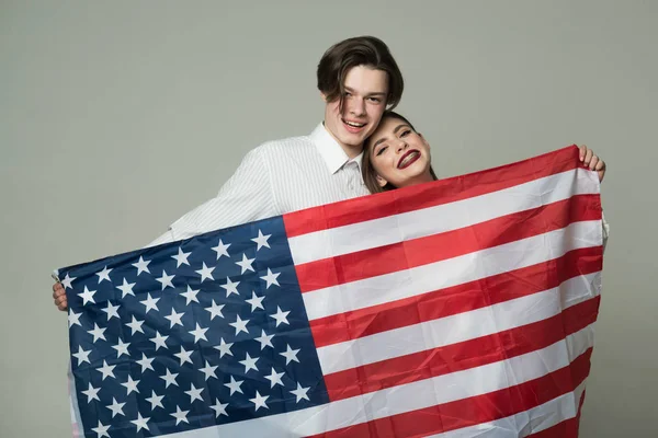 Sonriente pareja adolescente enamorada posando con la bandera de EE.UU. aislada sobre fondo gris. Feliz chico guapo en camisa blanca y chica bonita con frenillos celebrando el Día de la Independencia, 4 de julio — Foto de Stock