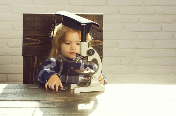 Criança inteligente em chapéu de graduação com estudo de microscópio no local de trabalho — Fotografia de Stock