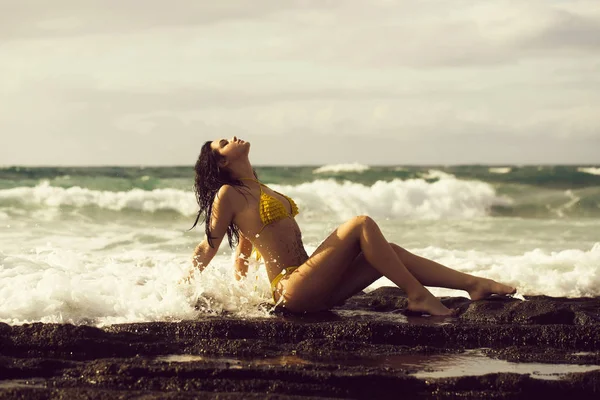 Bella ragazza in costume da bagno giallo sexy seduta sulla spiaggia rocciosa — Foto Stock