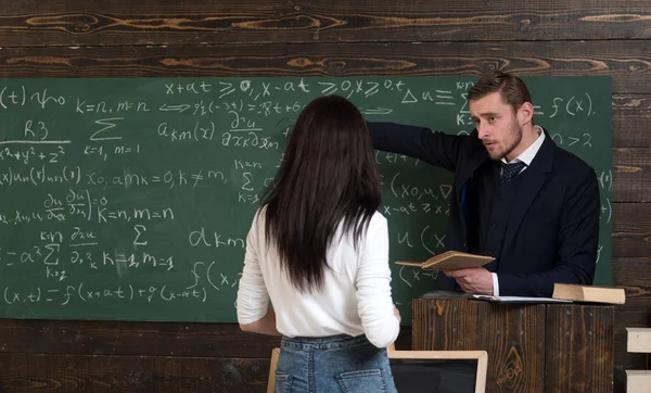 Учитель математики указывает на зеленую доску. Профессор дает объяснения брюнетке студентке. Поверните девушку в белом свитере, стоящую перед трибуной — стоковое фото