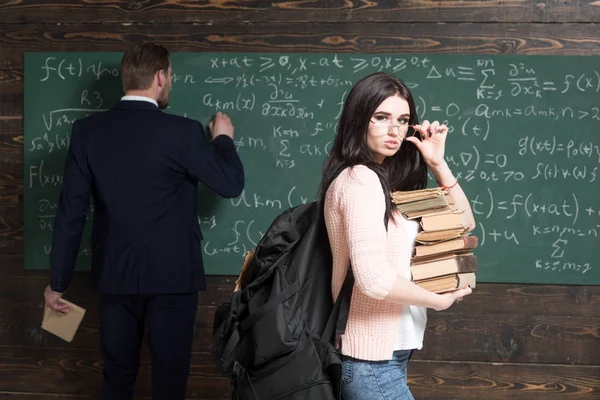 Studente bruna con mucchio di libri in piedi in classe mentre il professore sta scrivendo la formula su tavola verde — Foto Stock