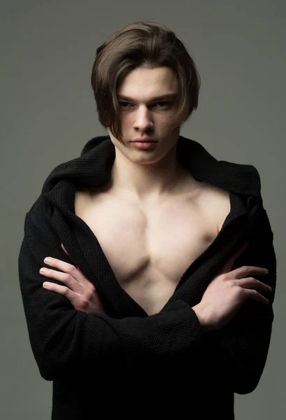 Hombre con la piel lisa en la cara, cuello y pecho mirando a la cámara. Chico con cara confiada en albornoz negro sostiene los brazos cruzados, fondo gris. Macho con corte de pelo largo con estilo. Concepto de masculinidad . — Foto de Stock