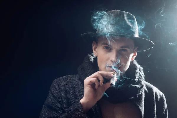 Hombre con abrigo, sombrero fumando cigarro, fondo oscuro. Macho en cara misteriosa, detective, investigador, agente. El tipo de traje anticuado parece misterioso con cigarro y humo. Concepto de detective Vintage . —  Fotos de Stock