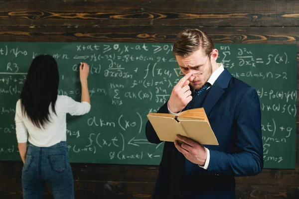 Vrouwelijke student vergelijking op het bord op te lossen. Jonge leraar met korte bristle aanpassen bril tijdens het lezen van een boo — Stockfoto