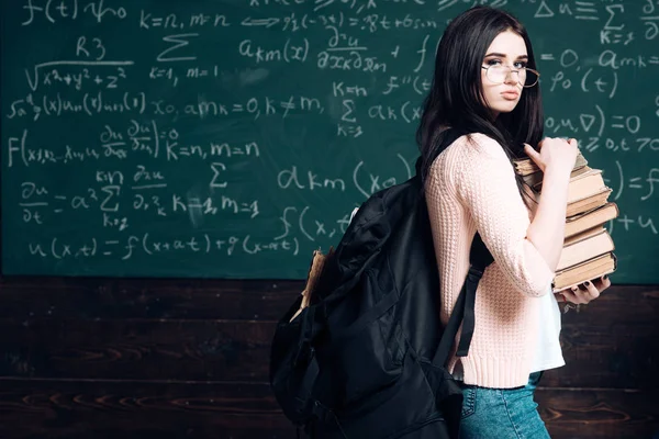 Chica morena de pie con una pila pesada de libros y una gran mochila negra frente a una pizarra verde llena de escritura. Preparación para el examen — Foto de Stock