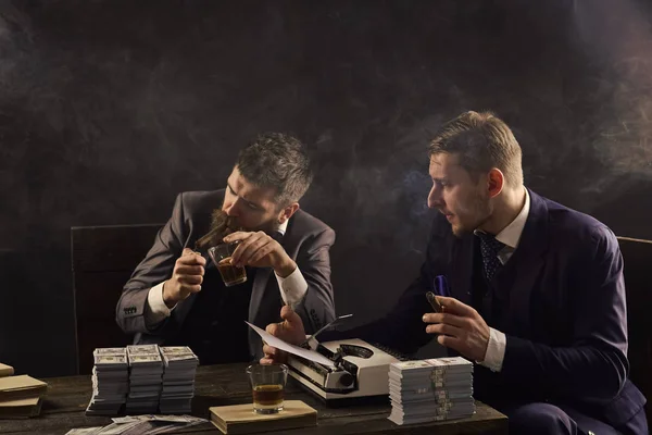 Hombres sentados a la mesa con montones de dinero y máquina de escribir. Concepto de negocio ilegal. Empresarios discutiendo trato ilegal mientras beben y fuman, fondo oscuro. Empresa dedicada a negocios ilegales . — Foto de Stock