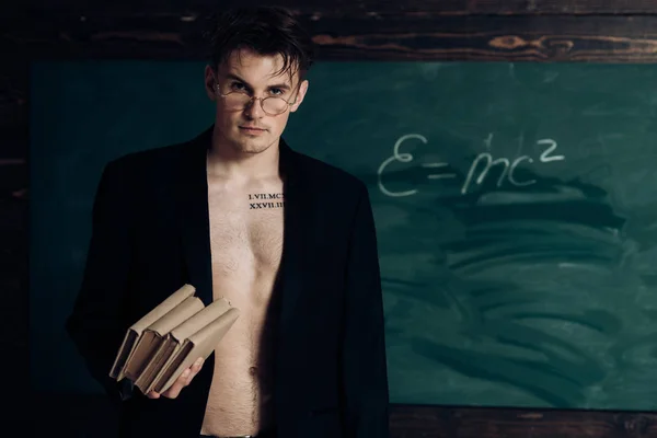 Teacher on pensive face, muscular figure, holds books in hand. Man with muscular torso, six packs, wears classic jacket and eyeglasses, looks attractive, chalkboard on background. Sexy teacher concept