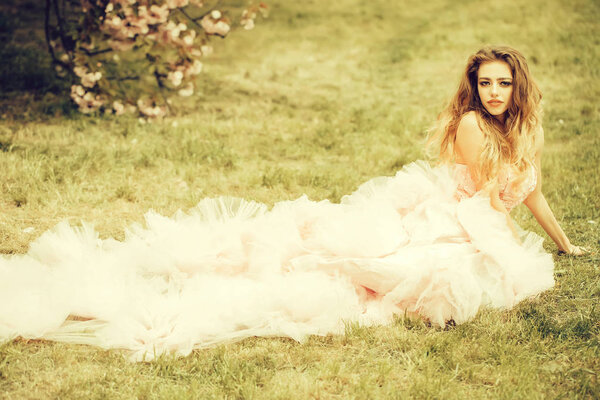 Beautiful young woman in pink glamour dress with long curly hair lying on green grass in spring sunny day
