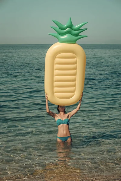 Woman holds above herself air mattress pineapple shaped, sea or ocean on background. Lady with air mattress stand in water, wearing stylish bikini and sunglasses. Summer vacation concept.