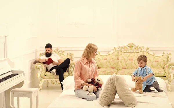 Father not pay attention to kid. Mother play with cute little son, while father busy with book. Indifferent father concept. Mom and son spend time together in bedroom, luxury interior background.