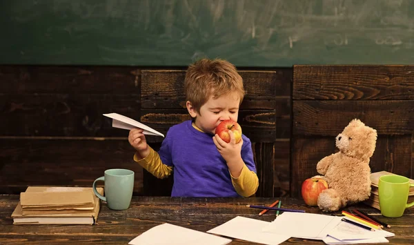 Ragazzo che mangia mela con piacere. Carino ragazzo mordere frutta con gli occhi chiusi. Bambino che gioca con aereo di carta — Foto Stock