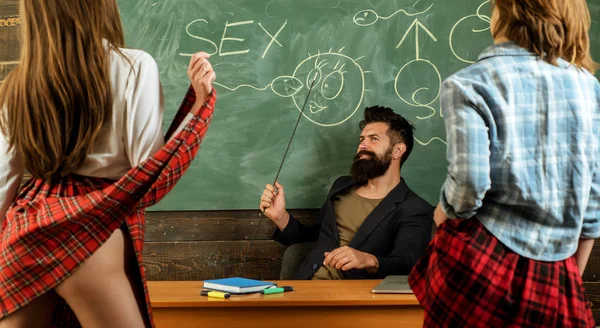 Sexy education. Bearded sexology teacher looks at two sexy female students. Erotic education and sex Symbols on chalkboard. Anatomy lesson and sex education in high school. Lets Talk Sex. — Stock Photo, Image