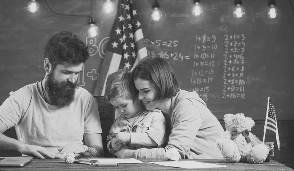 両親は息子に再生のアメリカの伝統を教えます。アメリカの国旗と教室、背景に黒板での両親との子供します。アメリカの家族は、息子とアメリカの旗と机に座ってください。愛国教育コンセプト. — ストック写真