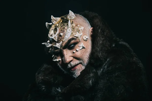 Alien, demon, sorcerer makeup. Horror and fantasy concept. Man with thorns or warts in fur coat. Demon on black background, copy space. Senior man with white beard dressed like monster in darkness.