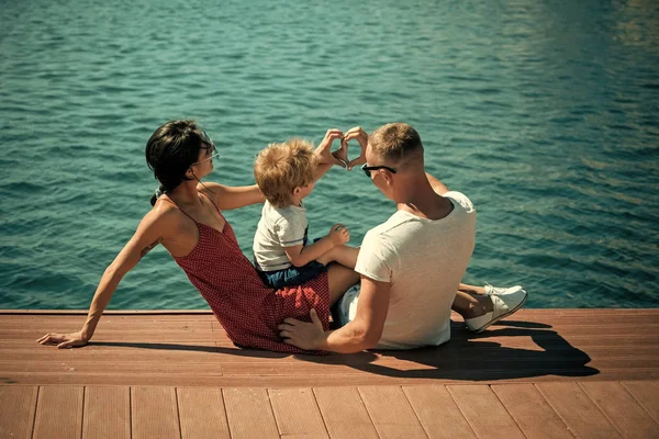 幸福的家庭一起度过时光, 海的背景。父母与儿子坐在海滨, 后景色。母亲和父亲在他们的孩子附近做心脏或爱手势与手。家庭度假概念. — 图库照片