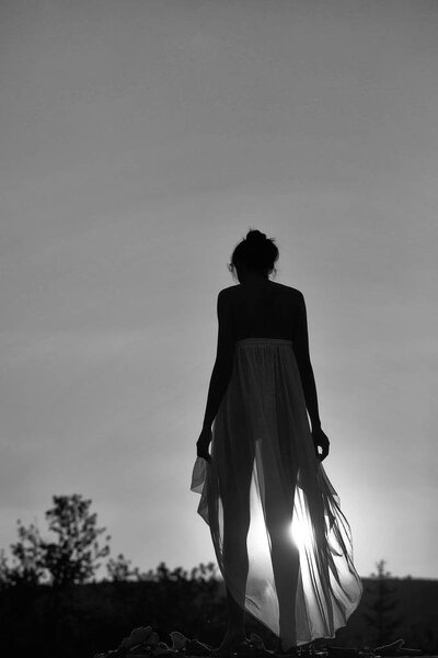 Silhouette of slim girl young woman female model in long transparent dress walks on nature over beautiful sunset