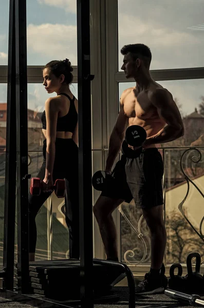 Man and woman in sportswear in gym, window on background. Couple does physical exercises with dumbbells together. Athletes, fitness trainers muscular and strong at gym. Sport and gym concept.