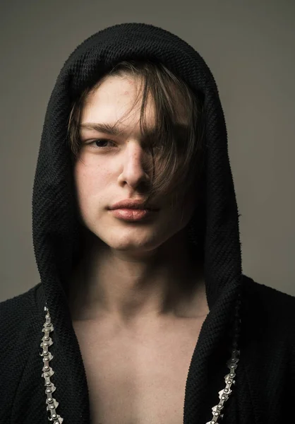 Beau garçon en capuche noire avec des cheveux noirs en couches de longueur moyenne avec une longue chaîne autour de son cou debout isolé sur fond gris, concept de mode jeunesse — Photo