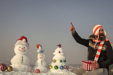 Mutlu bir adam mavi gökyüzü işaret parmağında. Sakallı hippi mevcut kutusuyla gülümseyen şapkalı. Kardan adam ve kar xmas ağacı kış gününde. Noel ve yeni yıl. Tatil kutlama kavramı