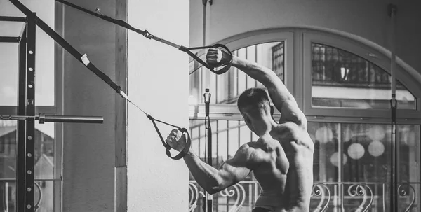 Sportman atleet, gespierde macho doet oefening met trx lussen, venster op de achtergrond. Man met naakte torso, en gespierde terug in sportschool geniet van training, trx, achteraanzicht. Sport en fitness concept. — Stockfoto