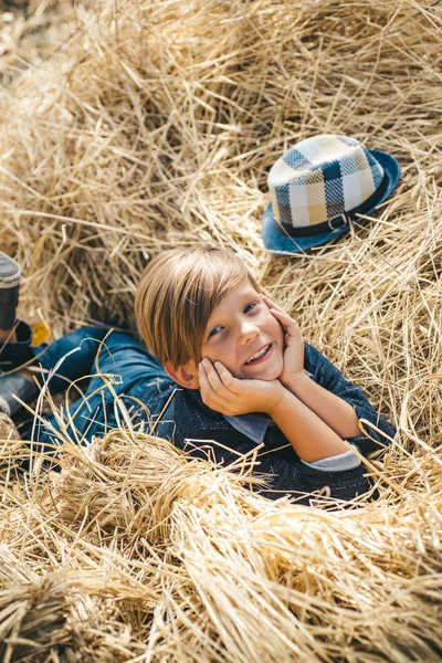 Lindo niño pequeño sosteniendo la hoja de oro en el fondo de la aldea granja. El niño yace en el heno. Lindo niño se están preparando para el día soleado de otoño . — Foto de Stock
