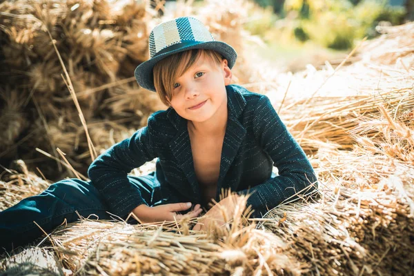 Söt liten barn pojke håller bladguld på gården byn bakgrund. Kid boy ligger på hö. Pojke i hatten förbereder för solig höstdag. — Stockfoto