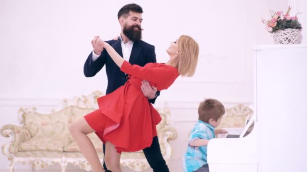 El chico toca el piano y los padres bailan. De ocio familiar. El hombre y la mujer barbudos vestidos de rojo bailan en la habitación blanca y el niño toca el piano. Pareja bailando . — Vídeos de Stock