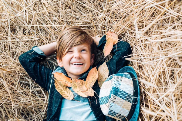Un ragazzino sorridente che gioca con le foglie e guarda la macchina fotografica. Il ragazzino giace sul fieno. Piccolo bambino carino che tiene foglia d'oro sullo sfondo del villaggio agricolo. Infanzia in fattoria . — Foto Stock