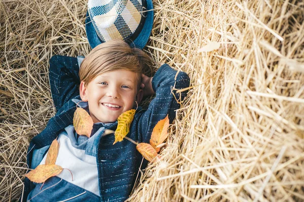 El niño yace en el heno. Lindo niño en el fondo de la aldea de granja. Un niño sonriente jugando con hojas y mirando a la cámara. Retrato de cerca. Buen tiempo en el pueblo . — Foto de Stock