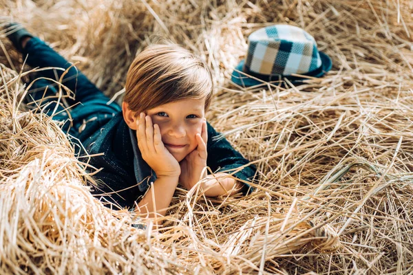 El niño yace en el heno. Lindo niño pequeño sosteniendo la hoja de oro en el fondo de la aldea granja. Niño en el sombrero se están preparando para el día soleado otoño. Infancia en la granja. Buen tiempo en el pueblo . — Foto de Stock