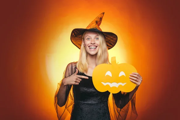 Surprised woman in witches hat and costume on red Halloween background. Attractive model girl in Halloween costume. Beautiful young surprised woman in witches hat pointing hand - showing products.
