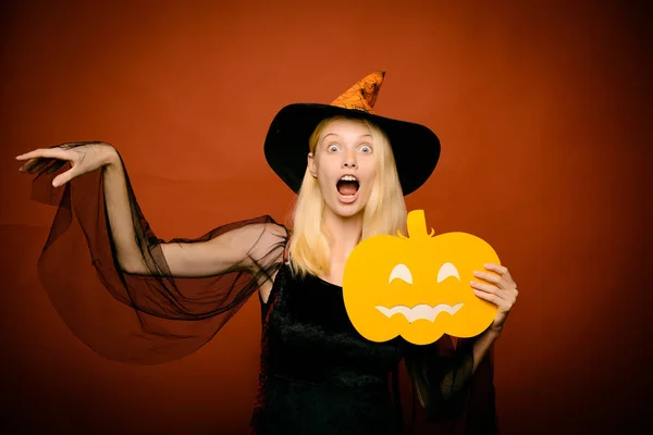 Surprised woman in witches hat and costume on red Halloween background. Pumpkin head jack lantern. Beautiful young surprised woman in witches hat pointing hand - showing products.
