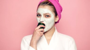Yüzünde bir maske ve havlu bir pembe bir salatalık yemek başında bir kız. Kadın giyen yüz maskesi. Yüz bakım kavramı.