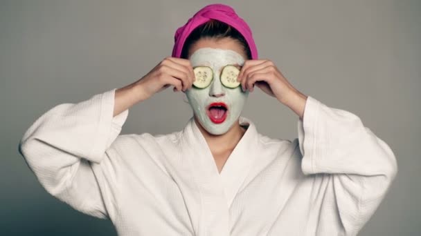 En tjej med en mask på ansiktet och en handduk på huvudet sluter ögonen med gurkor på en grå bakgrund. Kvinnan bär ansiktsmask. Face care koncept. — Stockvideo